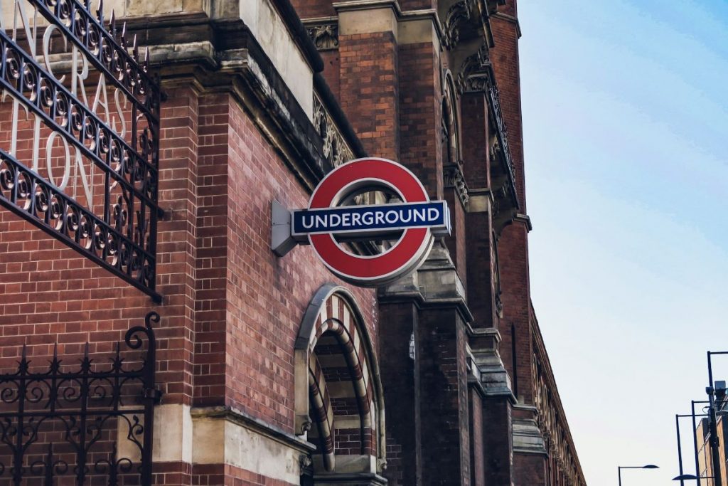 A bright red and blue London Underground sign marks an entrance on a brick building under a clear blue sky in central London.