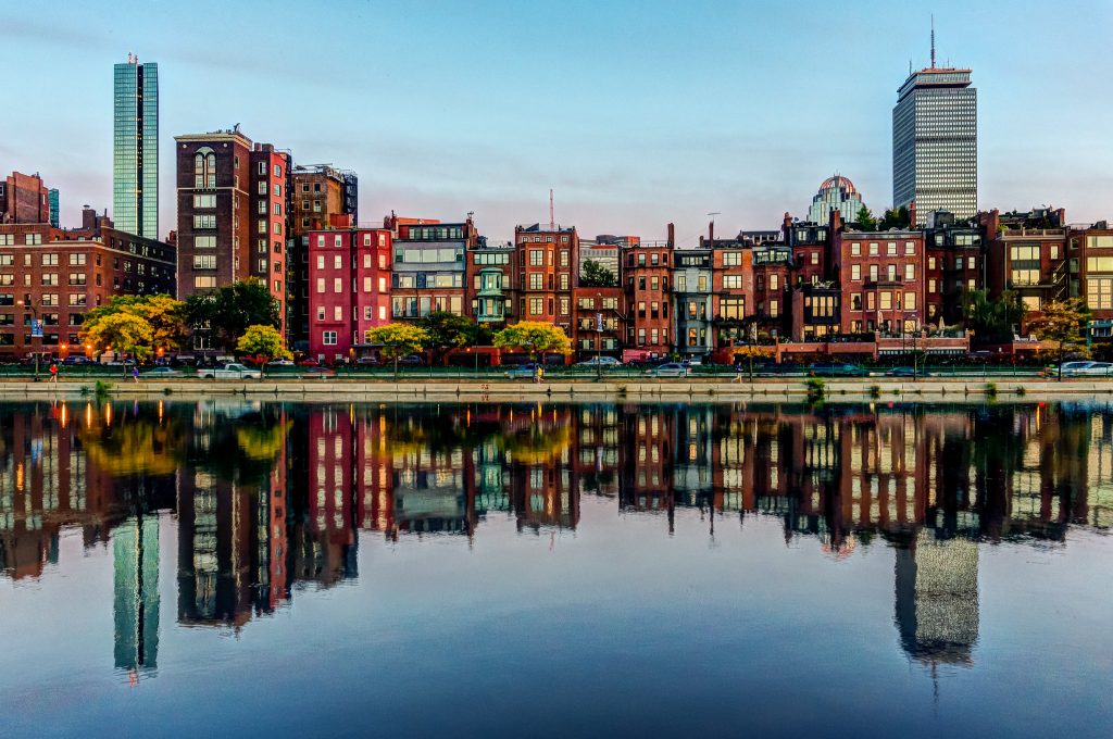 A row of brightly colored buildings and tall skyscrapers stands along Boston's waterfront, reflecting clearly in the calm harbor water.