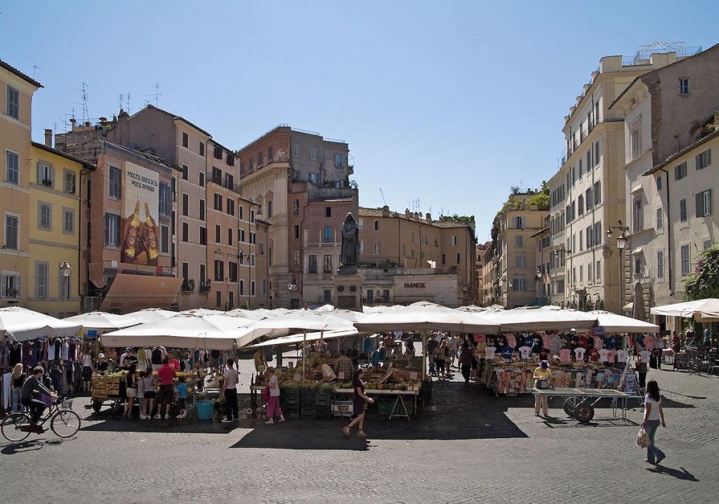 People shop and stroll among white-canopied stalls in a sunny Roman piazza surrounded by tall, historic buildings.
