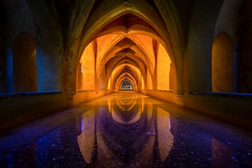 A long arched hallway in the Alcazar of Seville reflects yellow and blue light in a calm, shallow pool beneath.
