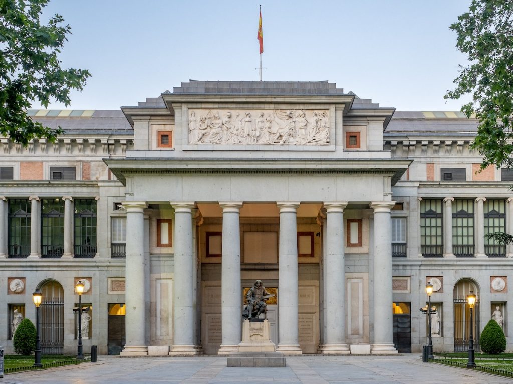 The Museo del Prado’s grand columned entrance in Madrid is adorned with carvings, a Velázquez statue, and the Spanish flag.