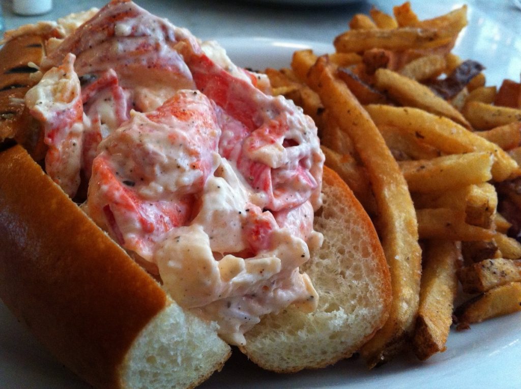 A lobster roll overflows with creamy lobster salad in a soft bun, paired with crispy French fries on a white plate.