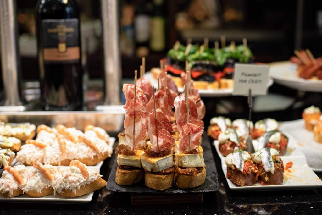 A selection of pintxos with ham, cheese, and vegetables sits on trays in San Sebastian Old Town, wine bottle behind.