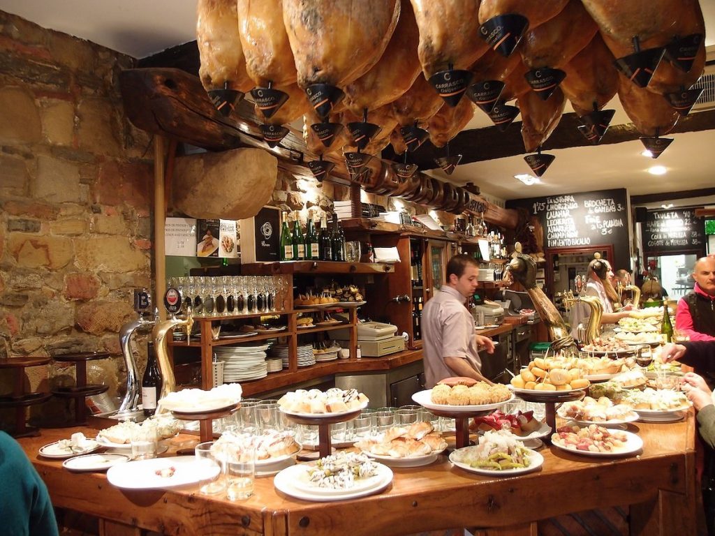Locals serve pintxos at a bustling tapas bar in San Sebastián, with hanging cured hams and shelves of wine bottles.