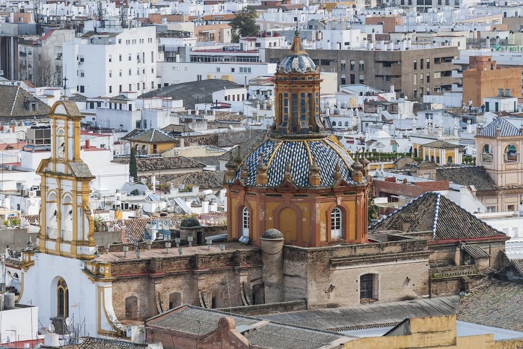 A historic church with a colorful tiled dome and bell tower rises above tightly packed white and beige buildings in Seville, Spain.