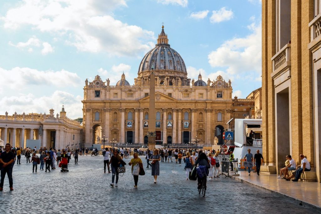 Large crowds gather in St. Peter’s Square at the Vatican, facing St. Peter’s Basilica and a tall central obelisk beneath clouds.