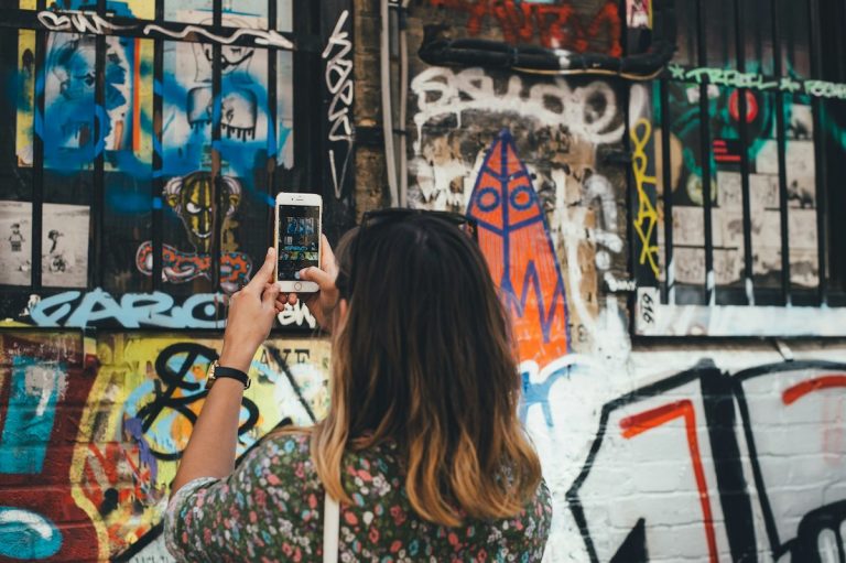A person in a floral shirt takes a picture of vibrant graffiti featuring abstract shapes and a rocket on a London street.