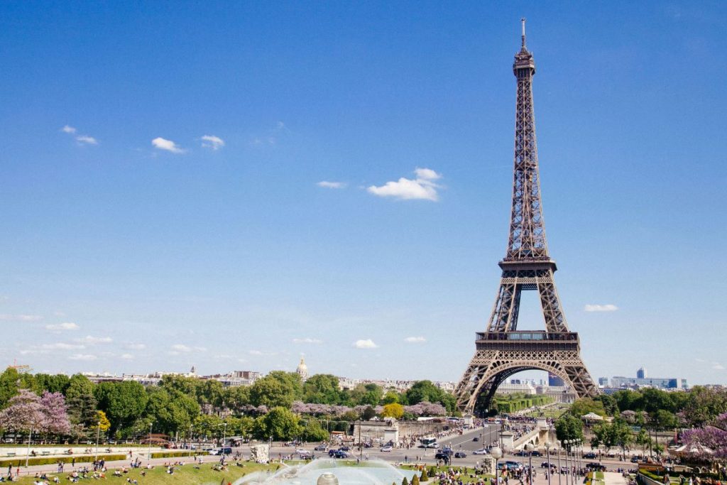 The Eiffel Tower rises above green gardens in Paris as people walk and relax, enjoying a sunny spring day.