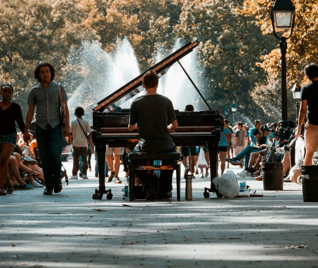 A man playing a piano in the middle of the park. People walk past him and watch him play live music.
