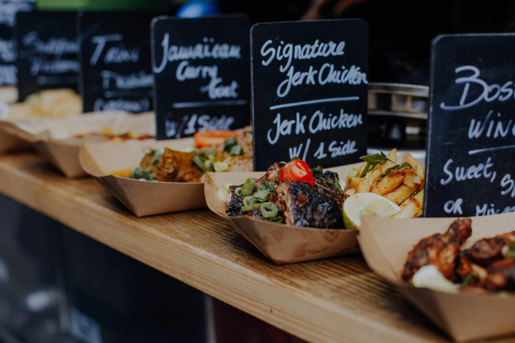 Several paper trays hold dishes like jerk chicken and curry goat on a wooden counter in a London food market. Handwritten blackboard menus are behind.
