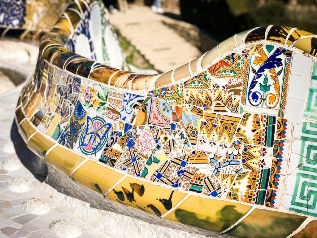 A detailed mosaic bench with vibrant tile patterns sits outdoors near stone steps, likely in Park Güell, Barcelona, by Antoni Gaudí.