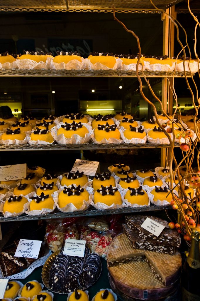 Rows of yellow pastries topped with chocolate and cream fill bakery shelves in Bergamo, surrounded by decorative branches and assorted sweets.