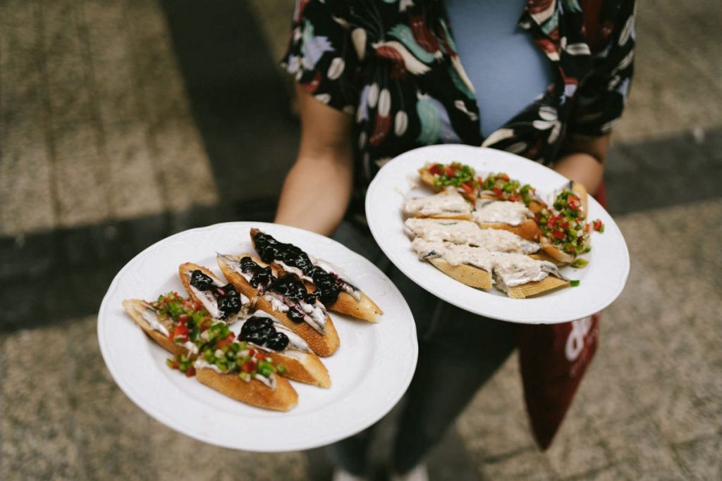 A person wearing a patterned shirt carries two plates of assorted pintxos with colorful toppings, standing on decorative tiles in San Sebastian.