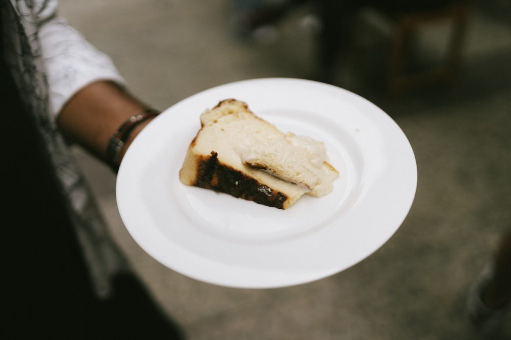 A hand holds a plate with bread pudding topped with creamy sauce, in San Sebastian Old Town, with blurred historic buildings behind.