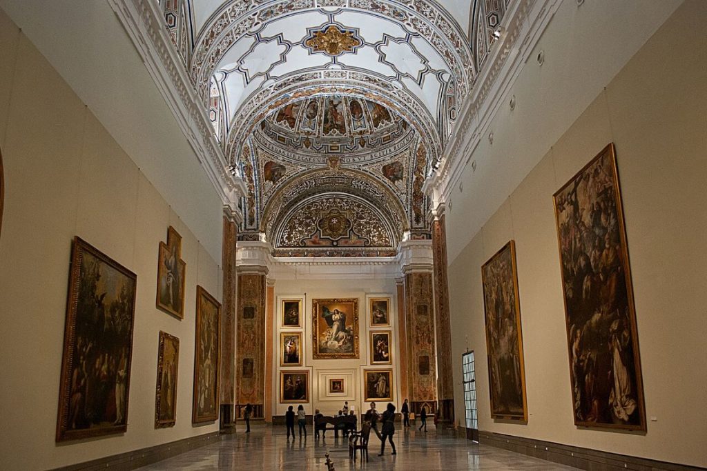 Visitors stand beneath a high, ornate vaulted ceiling, admiring large paintings in the spacious, well-lit Seville art gallery.