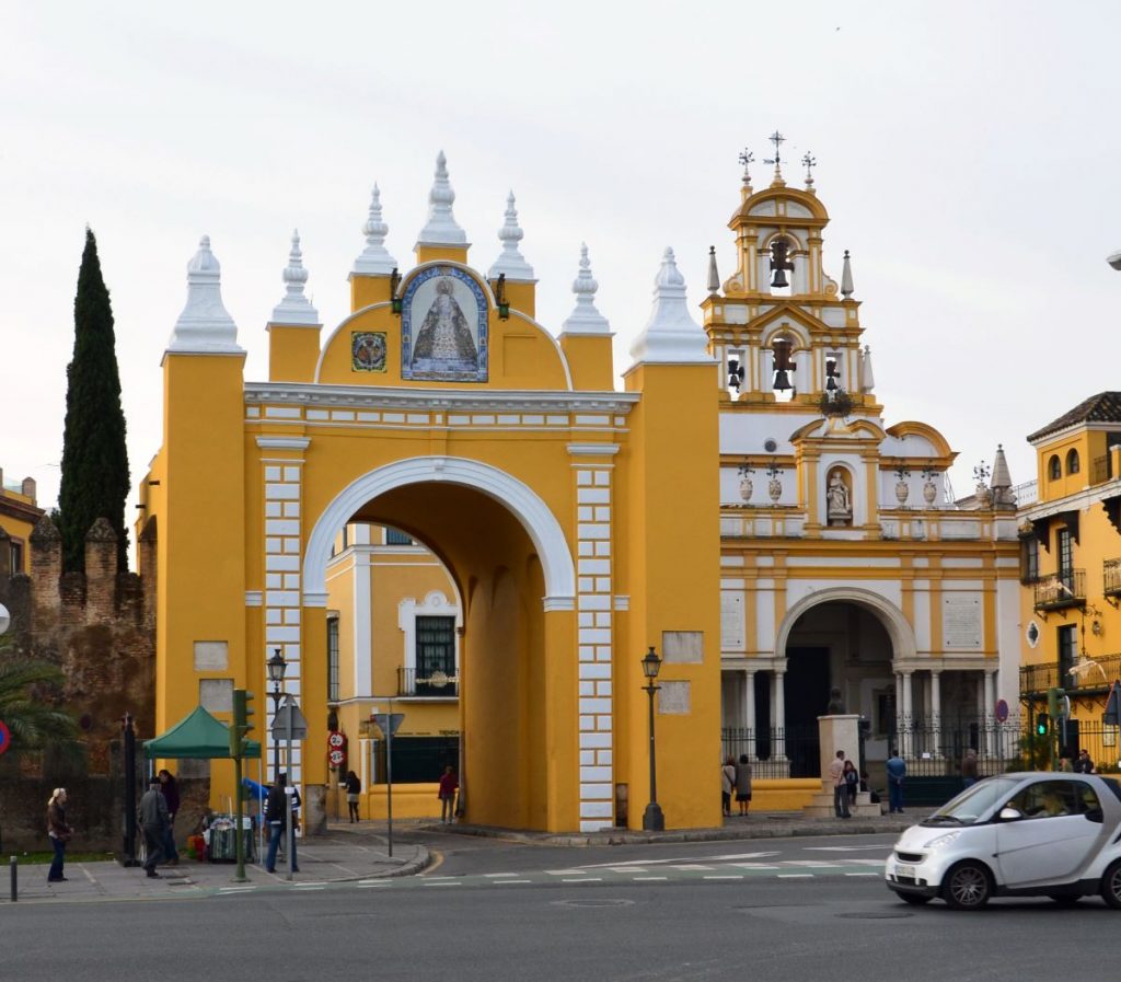 A yellow and white arched gateway stands beside Seville’s Iglesia de San Luis, with people and cyclists moving through the busy square.