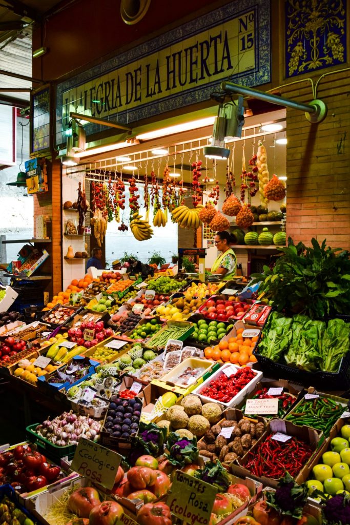 A market stall in Seville labeled “ALEGRÍA DE LA HUERTA” displays colorful fruits and vegetables, with a vendor standing behind the counter.