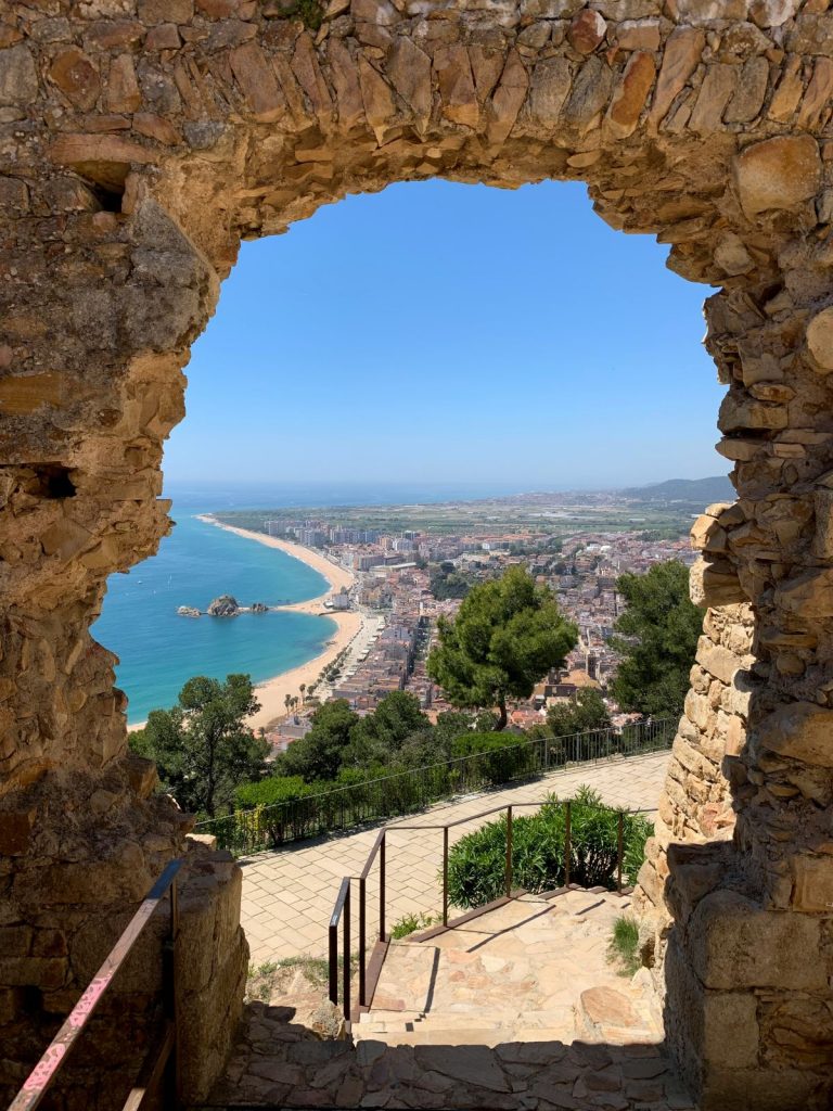 A large stone archway frames a sandy beach, clear blue sea, and coastal town near Barcelona under a bright sky.