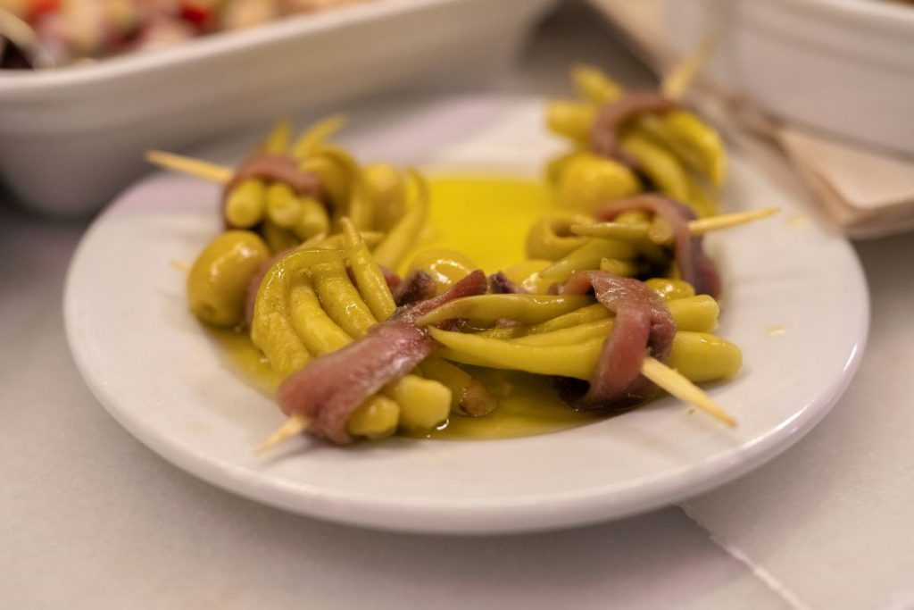 A white plate holds a Basque country classic, gildas, which are green chili peppers, olives, and anchovies on skewers with olive oil; napkins behind.