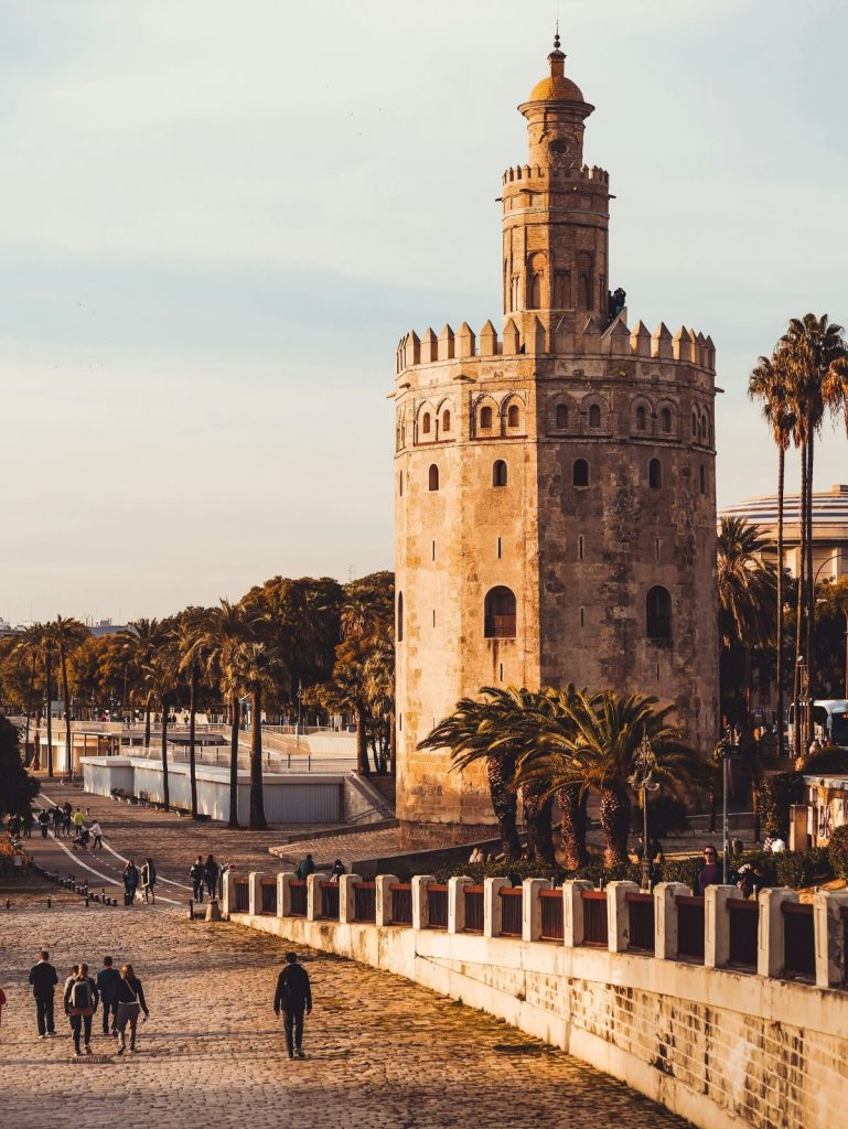 The Torre del Oro, a historic stone tower in Seville, overlooks the palm-lined riverwalk as people walk and cycle nearby.