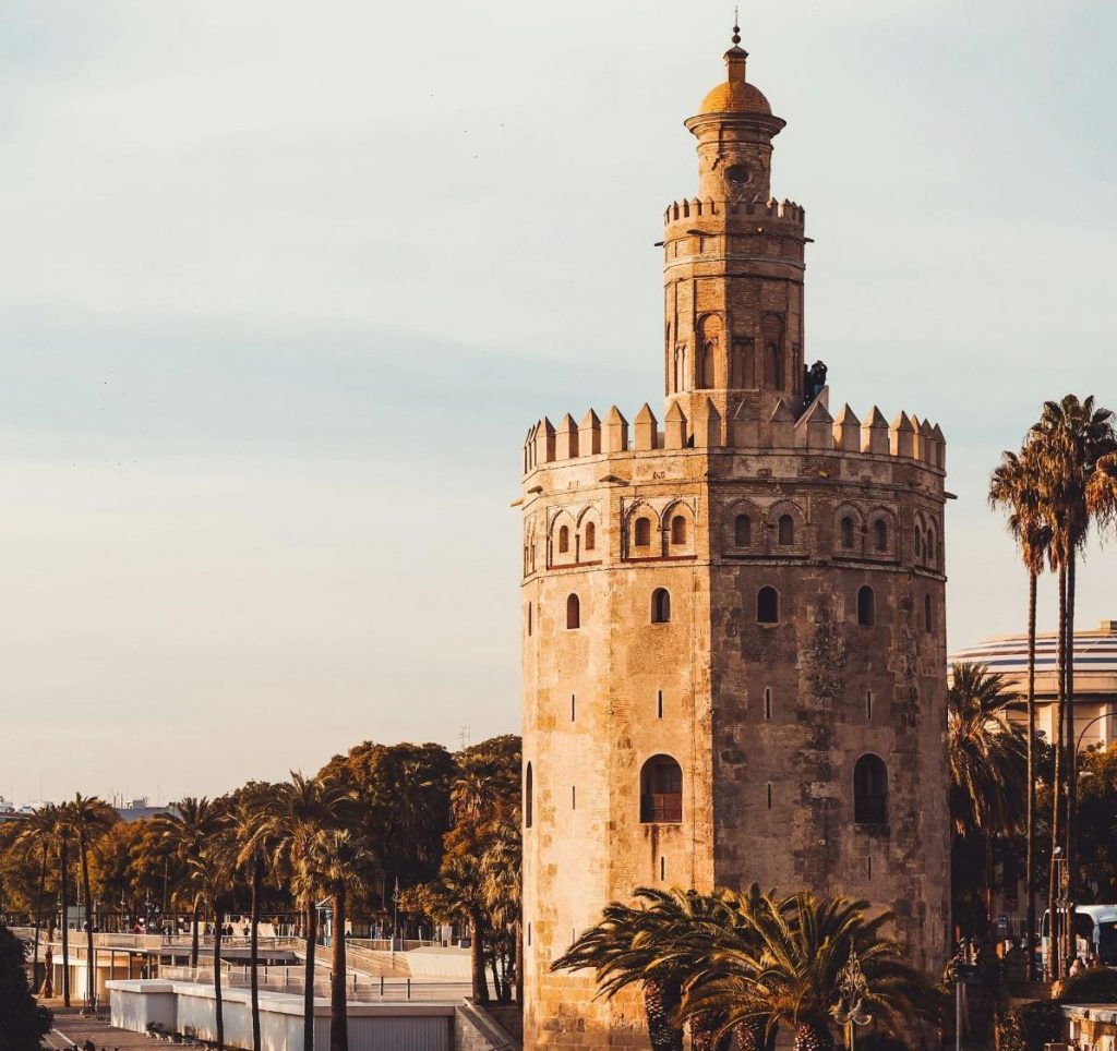 The Torre del Oro, a historic stone tower in Seville, overlooks the palm-lined riverwalk as people walk and cycle nearby.