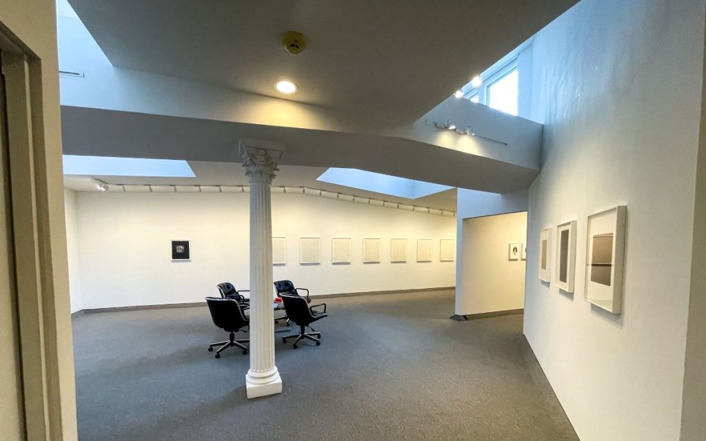 an interior of a minimalist art gallery exhibition, with white walls, and ceiling beams with skylights, and a pillar int he centre