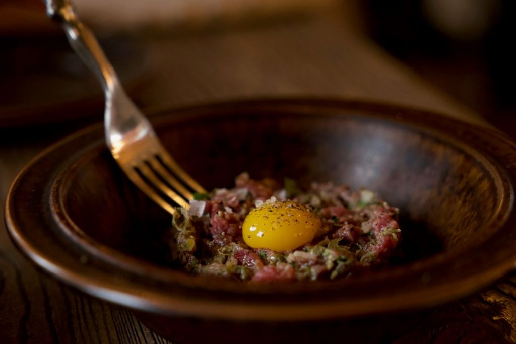 A person dipping their fork into a steak tartar with an egg over easy on top.