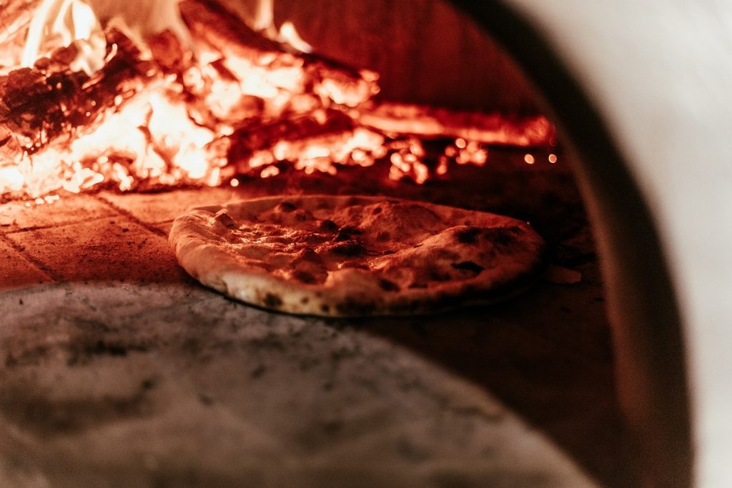 A Neapolitan pizza bakes in a wood-fired oven, flames curling around the crust in Sanità, Naples’ traditional pizzeria.