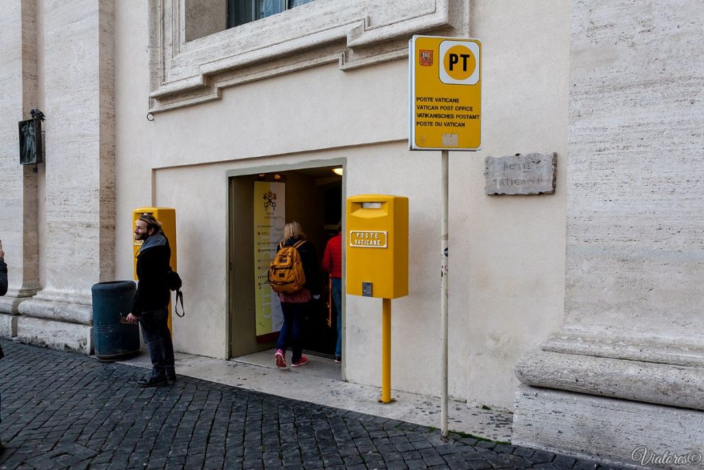 Several people approach the Vatican City post office entrance, passing a yellow mailbox and signs mounted on beige stone walls.