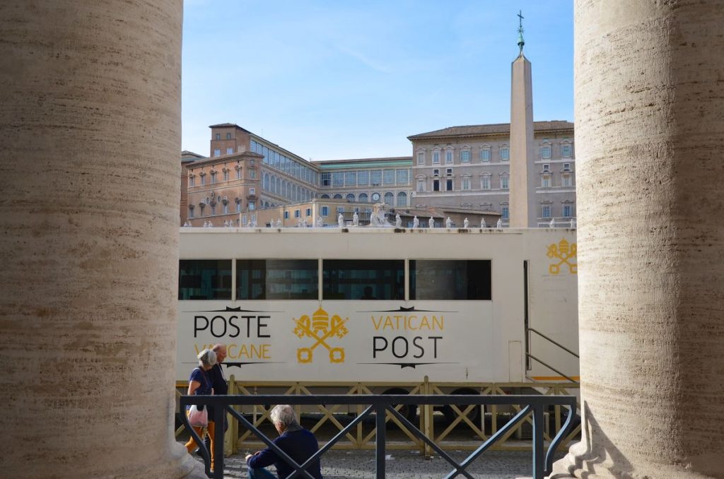 A yellow Vatican City post office trailer sits beside St. Peter’s Square columns, with visitors resting and the iconic obelisk visible.