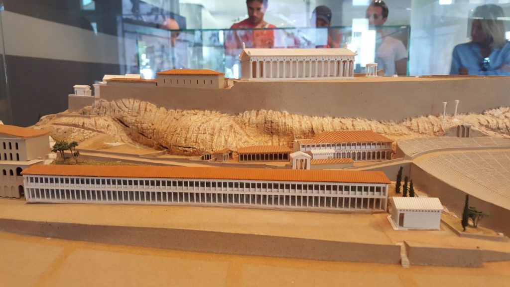 A 3D replica of the Acropolis in a glass display as people walk around in the background.