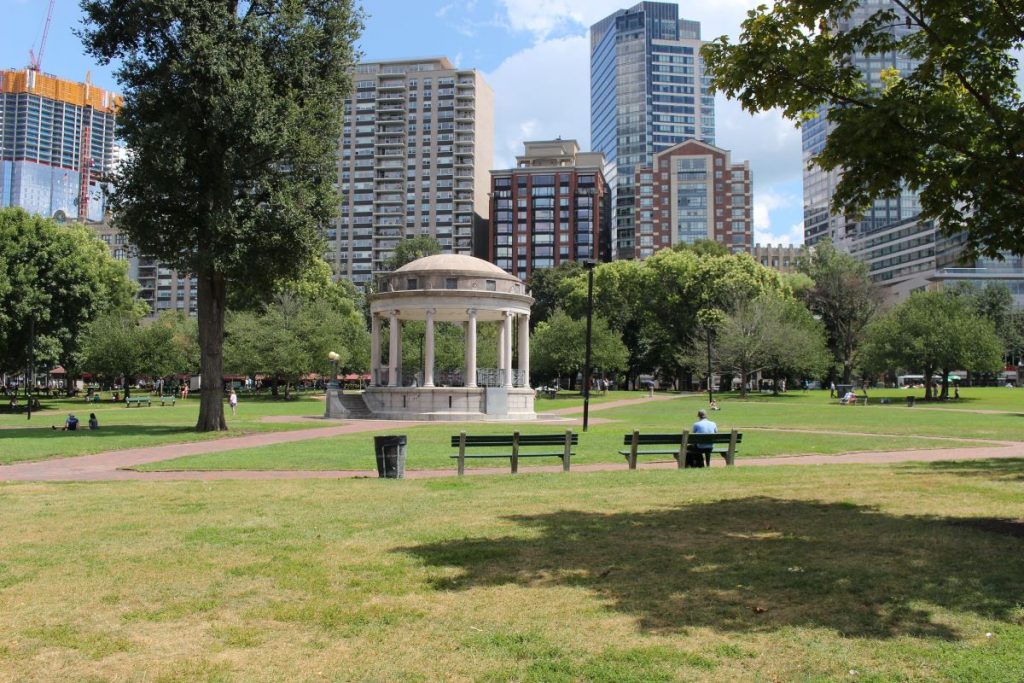 A round stone pavilion with columns sits in a green park in Boston, surrounded by people, benches, and city skyscrapers.