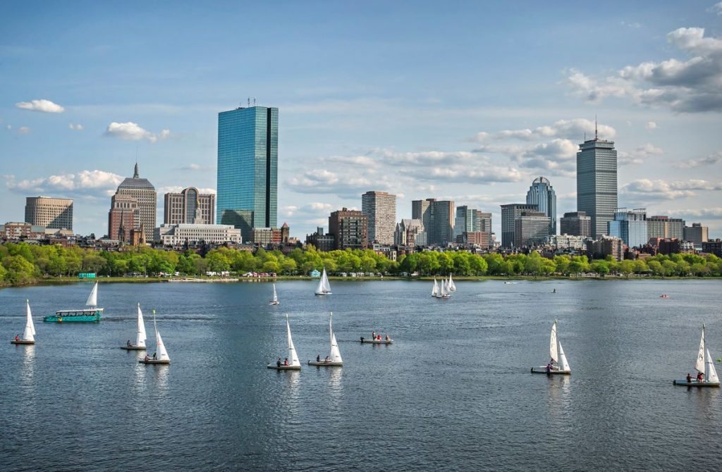 Several sailboats move along the Charles River in Boston, with tall glass skyscrapers and historic buildings rising behind leafy trees.