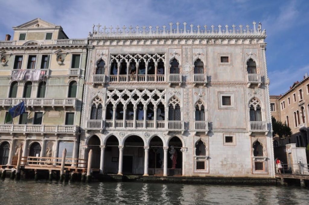 A grand Venetian Gothic palace with detailed arches and balconies stands beside a canal in Venice, Italy under a clear sky.