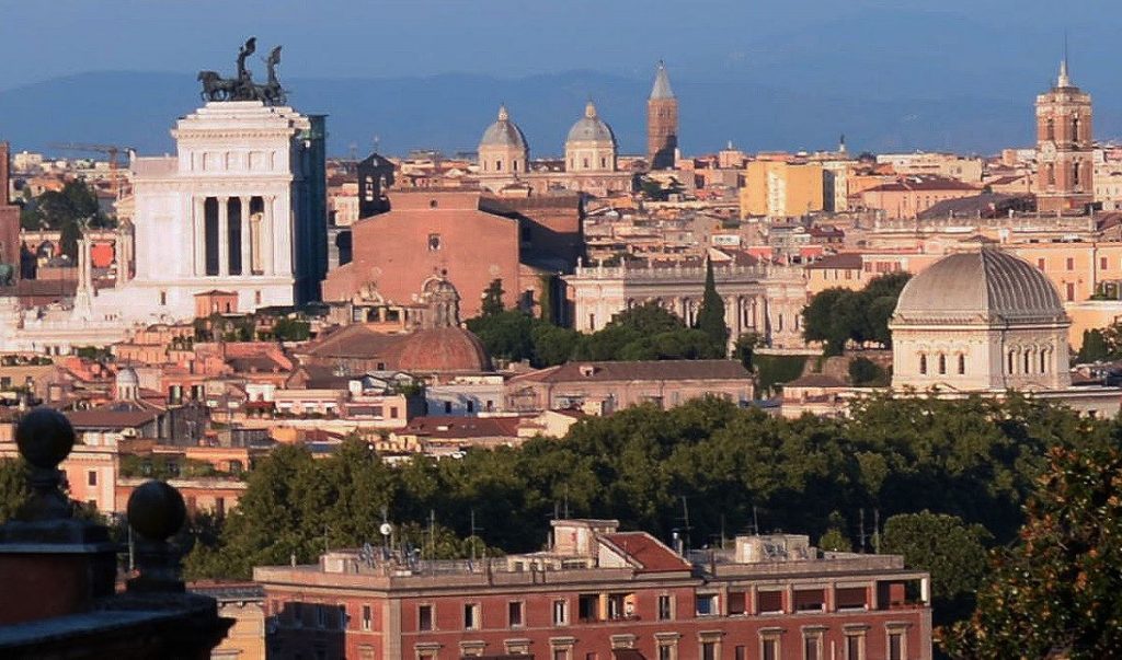A panoramic view of Rome shows the Vittoriano monument, historic domes, and Trastevere’s rooftops with trees beneath a clear sky. Tips for visiting Rome - go to the Giancolo Hill for panaromic views