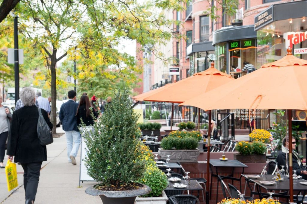 People walk past outdoor café tables with orange umbrellas, surrounded by flowers and brick shops on a sunny Boston street.