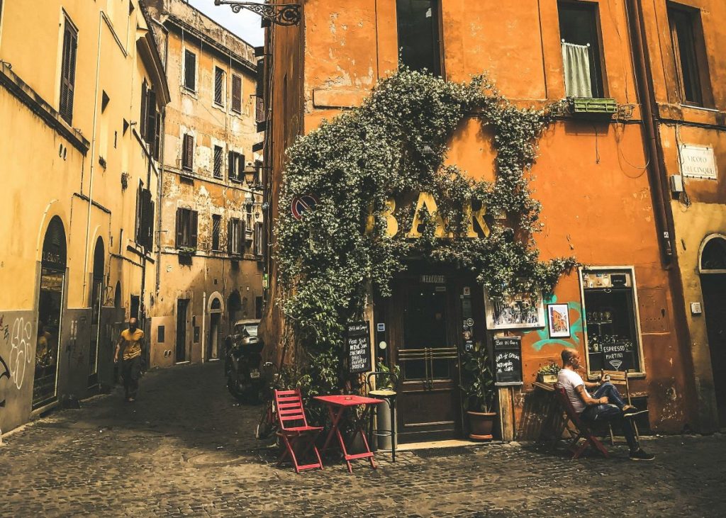 A person reads at a red table outside a vine-covered café and bar on a cobblestone corner in Trastevere, Rome. One of the tips for visiting Rome, is to enjoy exploring the small street aways from the big attractions