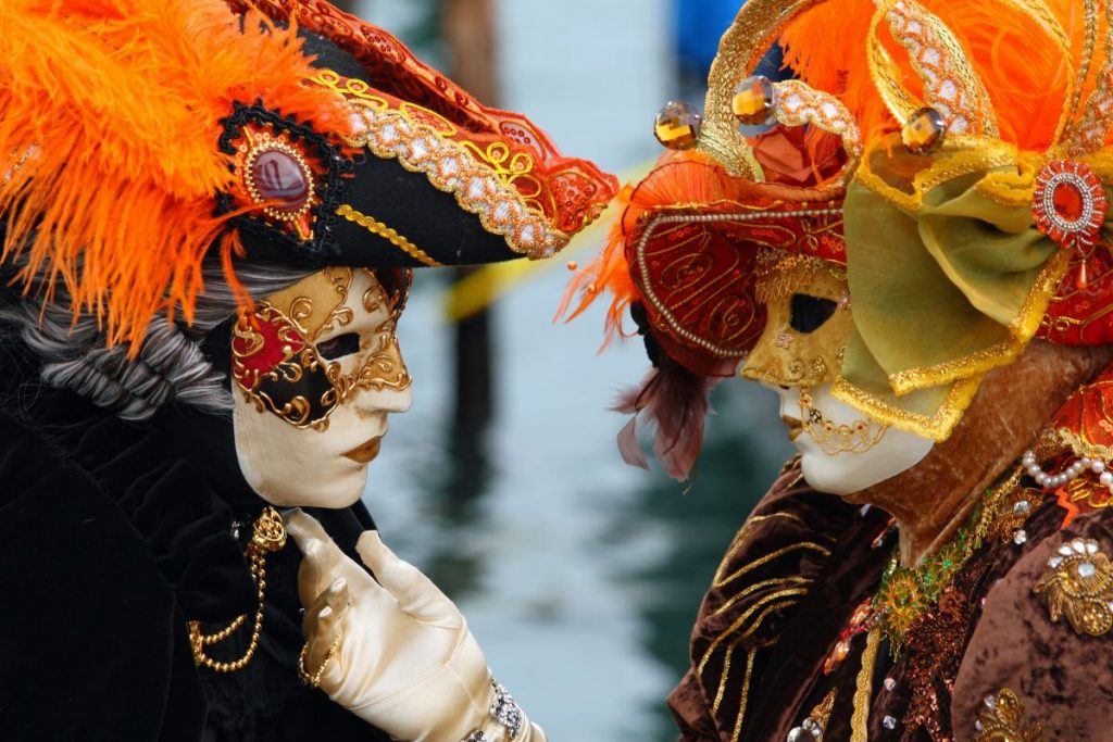 Two people in ornate Venetian carnival costumes and masks stand face to face in Venice, wearing feathered hats and gold embroidery.