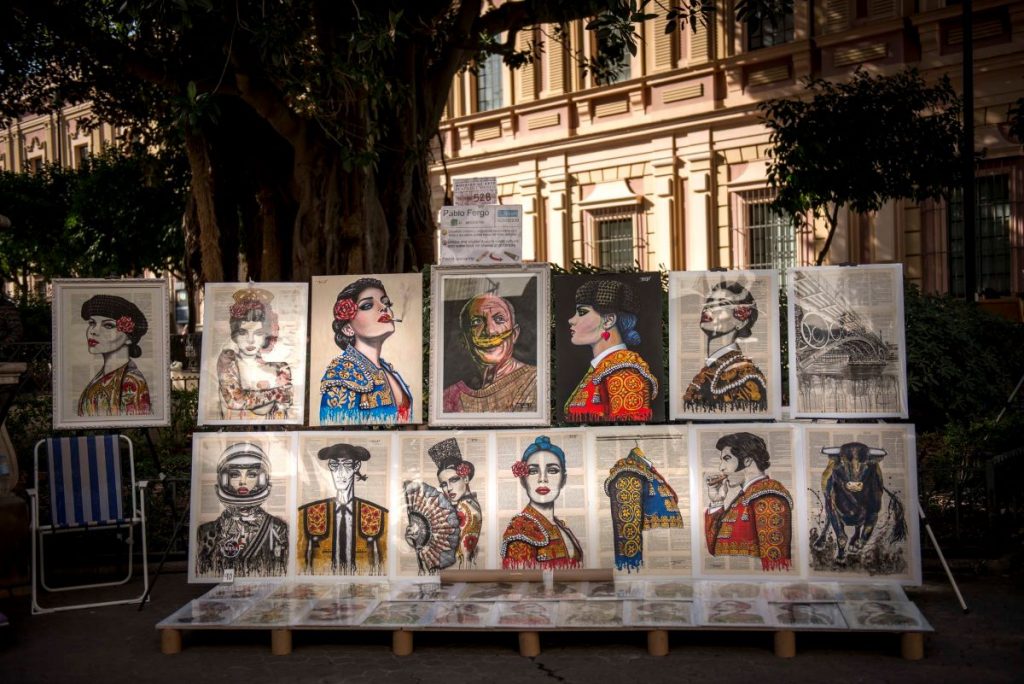 Colorful portraits of people in ornate clothing, including matadors, are arranged on easels in Triana, Seville near a historic building.
