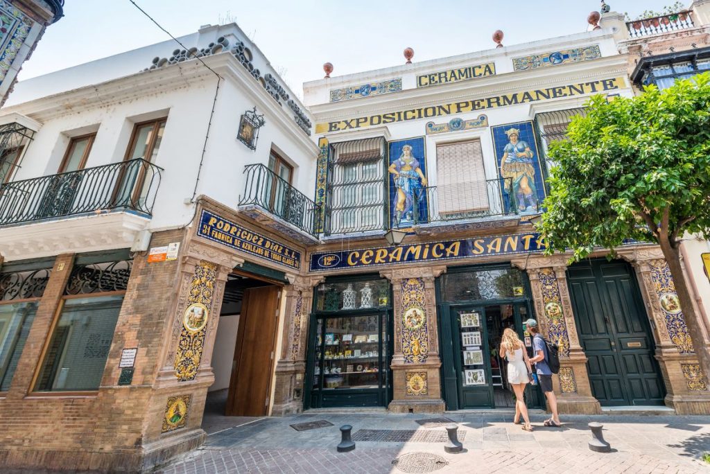 Two women stand outside Ceramica Santa Ana in Triana, Seville, surrounded by vibrant tiles and a sunny historic street.