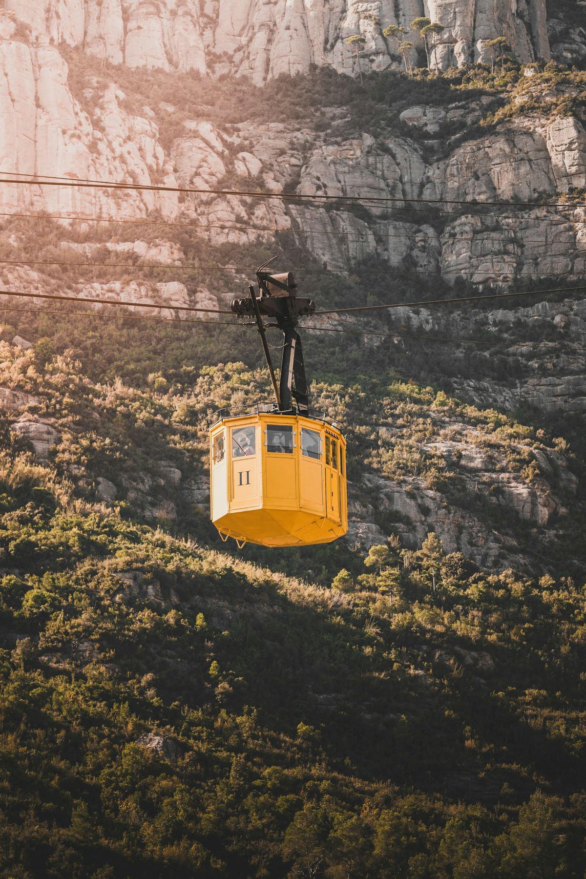 A yellow cable car labeled 11 glides above green trees and rocky mountains in Montserrat, Spain, under clear sunlight.