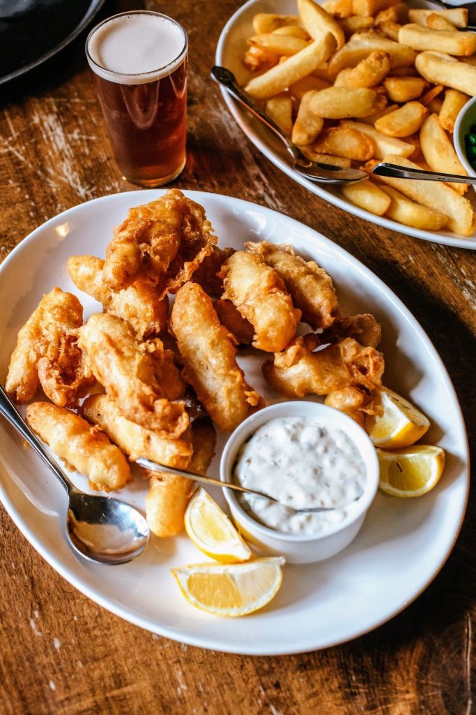 A plate of golden fried fish with tartar sauce, lemon wedges, thick fries, and beer sits on a London pub table.