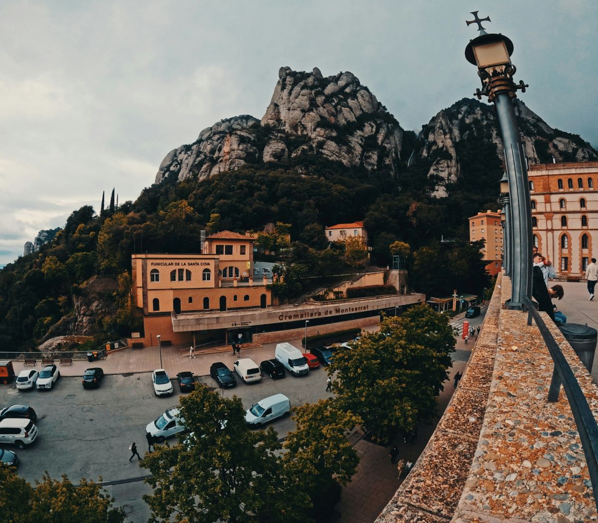 Rocky mountains of Montserrat, Spain rise behind green trees, yellow buildings, parked cars below, and a foreground stone terrace with lamppost.