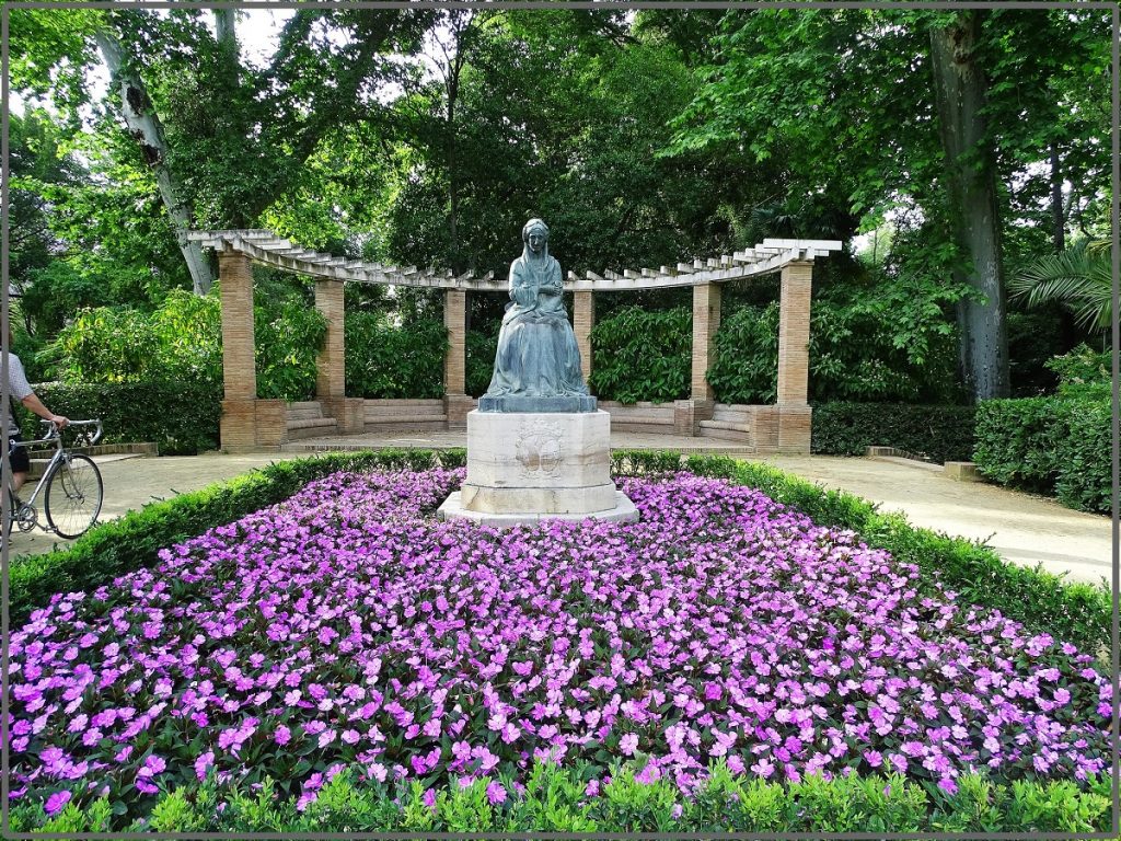 A bronze statue of a seated woman sits beneath a stone pergola, surrounded by lush greenery and blooming purple flowers in Seville.