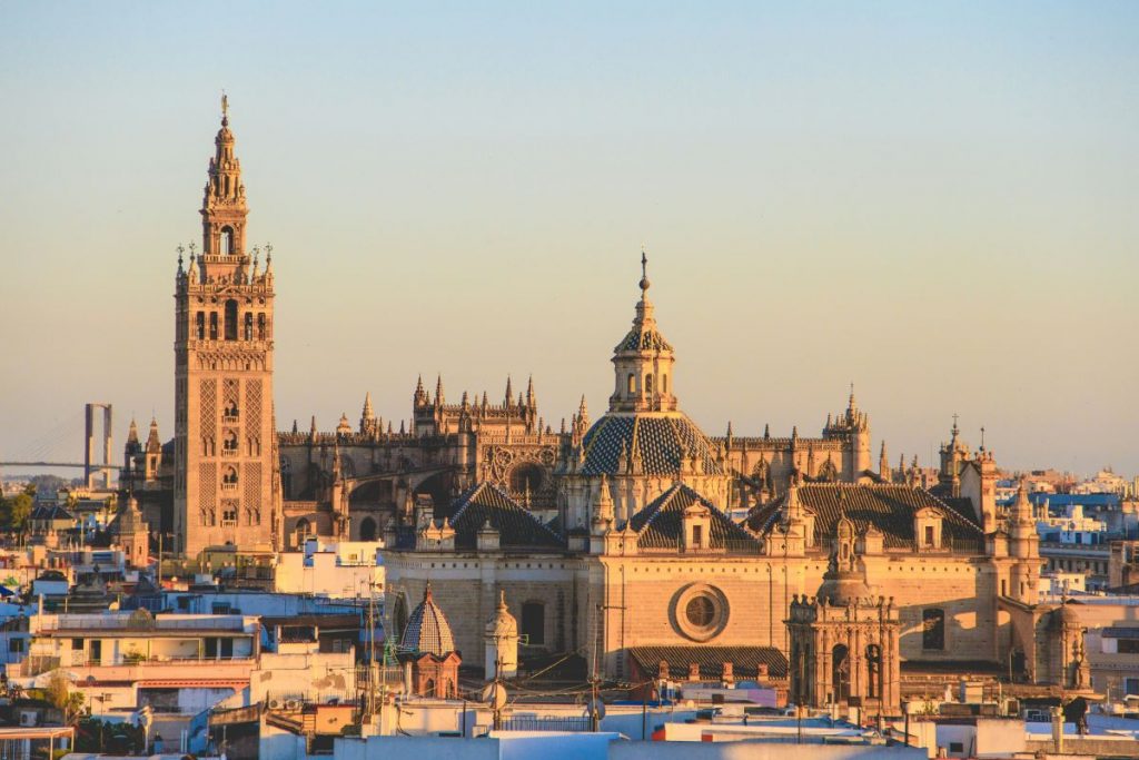 Seville Cathedral and the Giralda tower glow in warm evening sunlight, surrounded by Seville’s cityscape under a clear sky.
