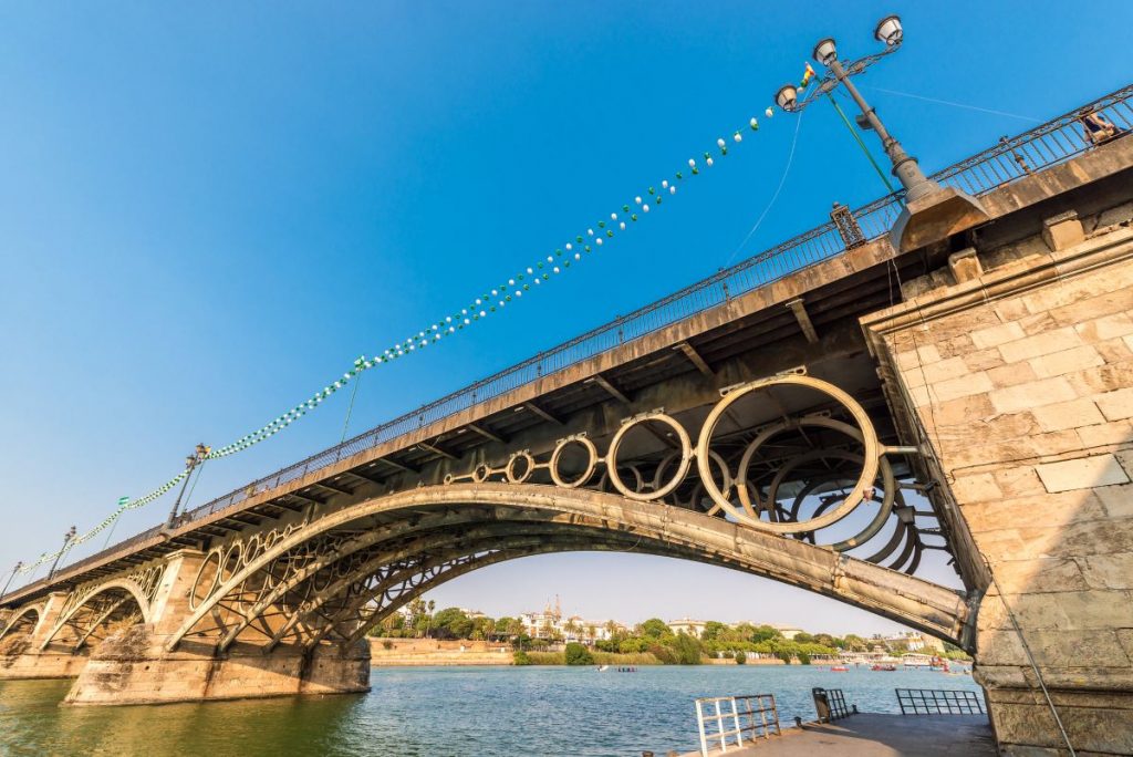 The Triana Bridge in Seville arches over the Guadalquivir River, featuring ornate metalwork and stone pillars, with city buildings behind.