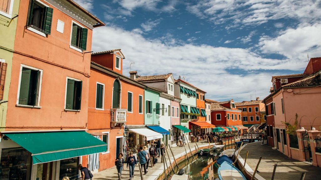 People stroll beside a canal with docked boats in Burano, Venice. Vibrant houses and green awnings line the sunlit walkway.