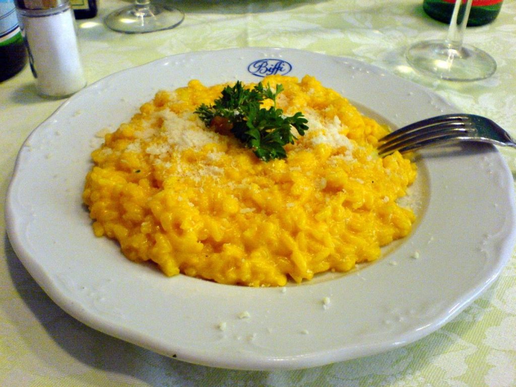 A famous milanese dish -  saffron risotto, a close up of a bowl of yellow risotto topped with parsley, in a white bowl on a restaurant table 