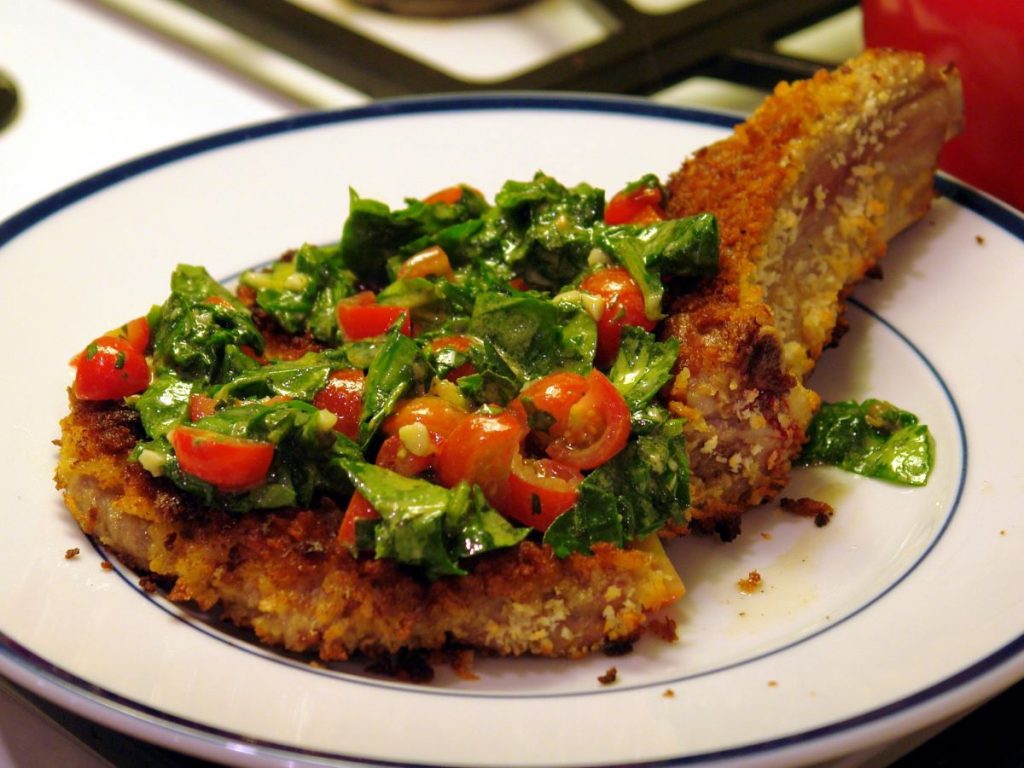 A close up photo of Cotoletta alla Milanese which is  a veal loin covered in breadcrumbs and fired, topped with a salad garnish of lettuce, tomato and pinenuts