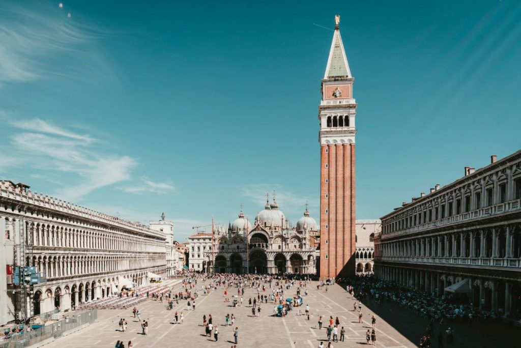 Crowds stroll through Venice’s St. Mark’s Square, with St. Mark’s Basilica centered and the Campanile bell tower rising nearby.